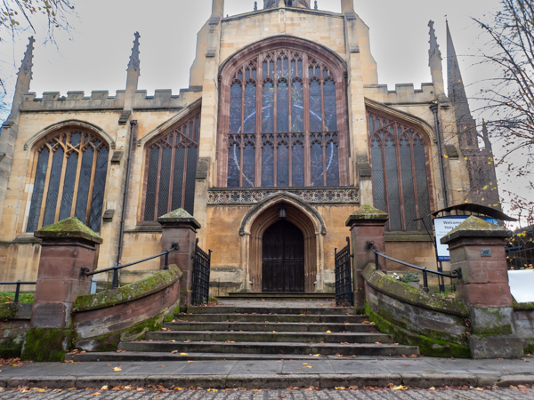 Colour photograph with a set of stone steps leading to a some open black gates.  Behind is a large black door with a pointed stone arch over it and a large stained glass window above the door with more stained glass windows on either side.  The church is made of yellow coloured stone with pinnacles on the roof.  The steps and the  wall at the edge of the steps are more red in colour.
